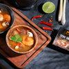 Tom Yum soup in a wooden bowl with ingredients on a dark surface, including a box of Siam Paolo Tom Yum soup.